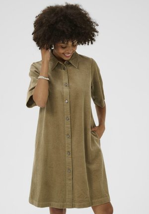 Femme avec des cheveux bouclés souriante, portant une robe en velours côtelé beige à boutons et à manches courtes, une main dans la poche, sur fond blanc.