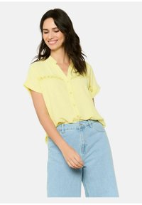 Blusa gialla con bottoni e colletto, caratterizzata da dettagli ricamati sul petto. Abbinata a jeans azzurri a vita alta.