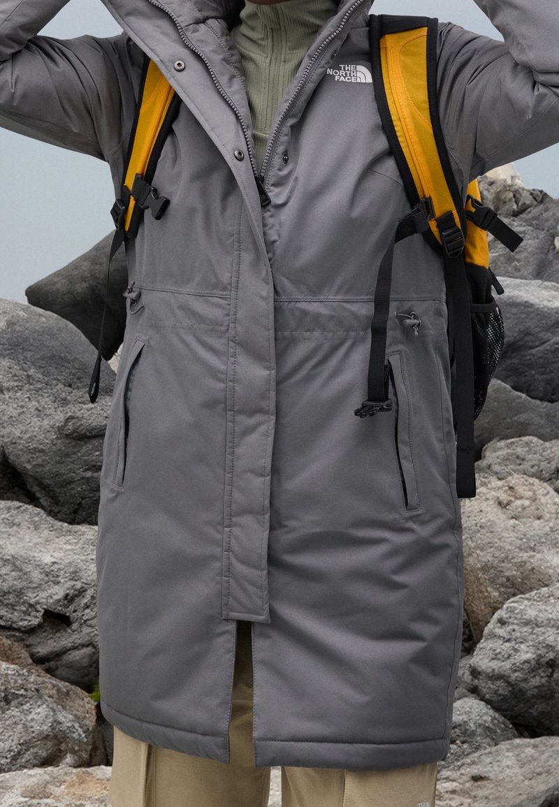 Personne portant un parka gris de North Face, avec des bretelles de sac à dos jaunes visibles, debout parmi de gros rochers gris en plein air.
