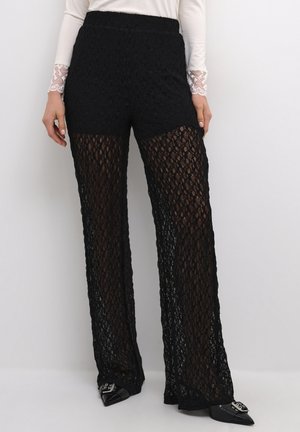 Femme portant un pantalon large en maille noire par-dessus un short noir, un haut blanc à manches longues avec des poignets en dentelle, et des chaussures noires pointues avec une boucle argentée.