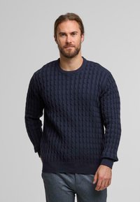 Maglione lavorato a maglia blu navy con un motivo a trecce testurizzato, scollatura rotonda e polsini e orlo a coste. Sono visibili dei pantaloni grigi abbinati.