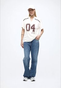 White jersey shirt with maroon numeral "04," paired with light blue flared jeans. Worn with a white cap and gray sneakers.