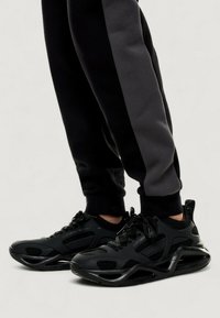 Zapatillas deportivas negras con un upper texturizado, paneles de malla y un diseño de suela acolchada. Combinadas con pantalones de chándal en negro y gris.