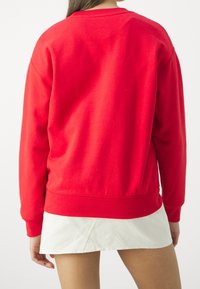 Personne portant un haut rouge vif à manches longues et une jupe courte blanche, vue de dos sur un fond uni.