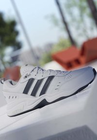 Baskets blanches Adidas avec trois bandes noires, lacets et semelle matelassée reposant sur une surface blanche en extérieur.