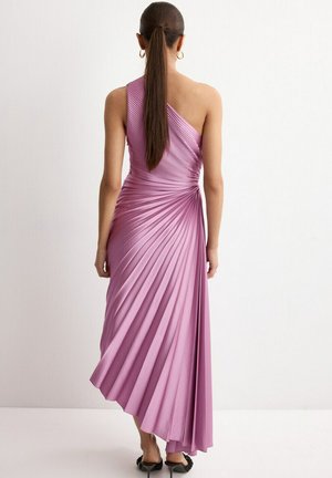 Femme aux longs cheveux foncés attachés en queue de cheval haute, portant une robe lilas plissée sans manches avec un ourlet asymétrique, debout contre un mur blanc.