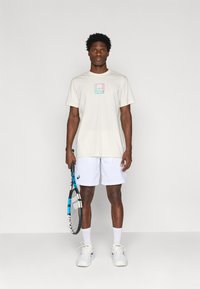 Κοντομάνικη κρέμα μπλούζα με τετράγωνο λογότυπο σε παστέλ χρώματα. Συνδυασμένη με λευκές σορτσάτες, λευκές κάλτσες και αθλητικά παπούτσια, κρατώντας μια ρακέτα τένις.