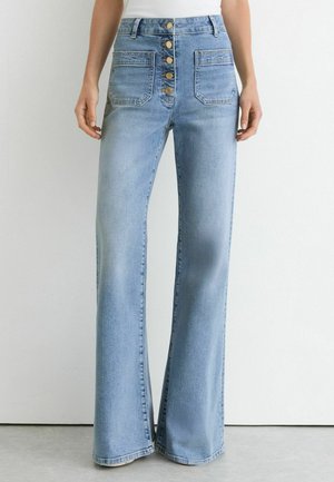 Kvinde iført lyseblå vidde jeans med høj talje, frontlukning med knapper og to store forlommer, stående mod en hvid væg.