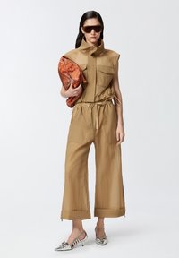 Bužna bezrukavna kombinezona s dva prednja džepa, elastičnom strukom i širokim skraćenim nogavicama. Drži narandžastu torbicu s uzorkom i nosi naočale za sunce.