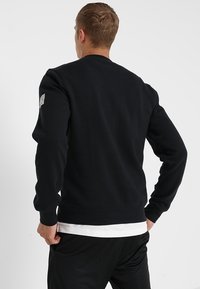 Uomo con capelli biondi corti che indossa una felpa nera sopra una camicia bianca e pantaloni neri, girato di spalle con le mani sui fianchi.