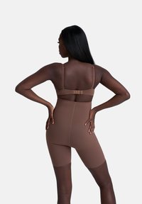 Vrouw met een donkere huid en lang steil haar staat met haar handen op haar heupen, gekleed in bruine high-waisted shapewear shorts en een bijpassende bh, van achter bekeken.