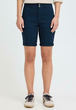 Kvinne iført marineblå shorts med mansjetter, beige genser og beige joggesko med svarte lisser, stående mot ensfarget lyst bakgrunn.