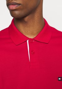 Polo rouge en tissu coton texturé, avec une patte de boutonnage à deux boutons, des accents blancs et un petit logo brodé sur la poitrine.