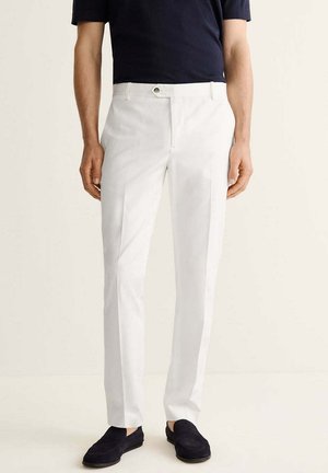 Hombre con pantalones blancos a medida con bolsillos laterales, mocasines negros y una camisa oscura de manga corta, posando sobre un fondo liso.