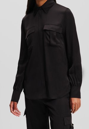 Femme portant une chemise noire à manches longues avec deux poches à rabat sur la poitrine et un pantalon noir avec une poche latérale.