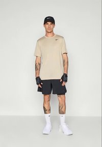 Homme debout portant un T-shirt beige Nike, un short noir Nike, une casquette noire, des gants sans doigts noirs, des chaussettes blanches Nike et des baskets blanches.