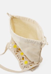 Beige tas met een trekkoordsluiting, gekleed met witte haakwerk madeliefjes met gele hartjes aan de basis. Binnenin is de tas van eenvoudig beige stof.