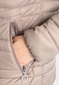 Lichtbruine pufferjack met een ritszak en zilverkleurige hardware. Gequilt ontwerp met een gladde textuur en aansluitende mouwen.