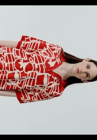 Femme aux longs cheveux bruns portant un tricot à manches courtes rouge et blanc à motifs avec des boutons, sur un fond clair uni.