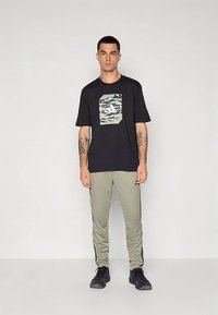 Μαύρο μπλουζάκι με τσέπη σε ύφος παραλλαγής στο μπροστινό μέρος, σε συνδυασμό με ανοιχτό πράσινο αθλητικό παντελόνι με μαύρες ρίγες στο πλάι. Μαύρα αθλητικά παπούτσια.