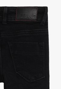 Černé džíny z denimu, pohled zezadu, ukazující pravou kapsu a černou koženou brandovou nášivku s červeným prošíváním na pasu.