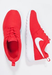 Červené sportovní boty z mřížkového materiálu s bílými akcenty, zaoblenou špičkou, šněrovacím designem a strukturovanou podrážkou. Očko na patě pro snadné obouvání.