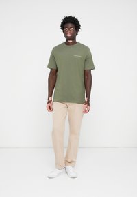 T-shirt en coton vert avec un col rond et des manches courtes, associé à un pantalon beige à jambe droite et des baskets blanches. Logo texte simple sur la poitrine.