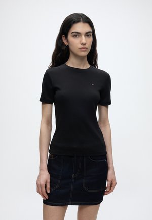 Νεαρή γυναίκα με μακριά σκουρόχρωμα μαλλιά, φορώντας απλό μαύρο κοντομάνικο μπλουζάκι και σκούρα τζιν μίνι φούστα, στέκεται μπροστά σε απλό φωτεινό φόντο.