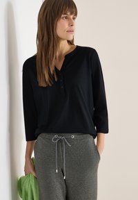 Femme portant un t-shirt henley noir et un pantalon gris à cordon, tenant un pull en maille vert, debout devant un mur clair uni.