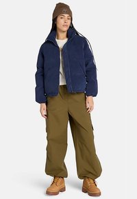 Chaqueta de pana azul marino con cuello alto, combinada con pantalones cargo oliva y botas marrones con cordones. Gorro marrón con detalle de logotipo.