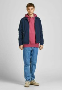 Marineblauwe jas met een rits, gelaagd over een roze hoodie. Lichtblauwe jeans en beige schoenen maken de outfit compleet. Het model staat rechtop.