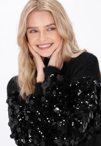 Pulover negru cu mâneci texturate și decorate cu paiete cu forme rotunde. Modelul are părul lung, ondulat, de culoare blondă și un zâmbet blând, având mâinile pe față.