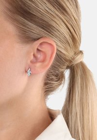 Närbild av en kvinnas öra med en silverfärgad, bladformad studs och blont hår som är uppsatt i en låg hästsvans.