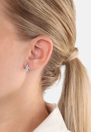 Primo piano dell'orecchio di una donna con un orecchino a forma di foglia in argento e capelli biondi legati in una coda di cavallo bassa.