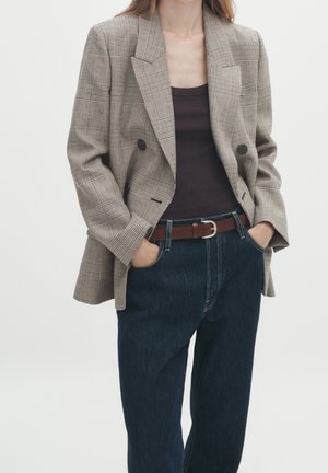 Woman wearing brown checkered double-breasted blazer over dark brown top, tucked into dark blue jeans with a brown belt, hands in pockets.