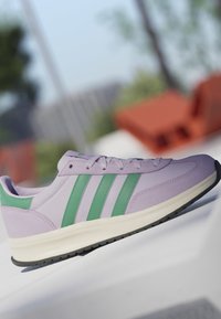 Zapatilla deportiva de color púrpura claro con rayas verdes y suela blanca colocada sobre una superficie blanca con un fondo exterior desenfocado.