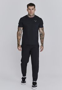 Schwarzes T-Shirt mit kurzen Ärmeln und einem Logo auf der Brust, kombiniert mit schwarzen Jogginghosen und schwarz-weißen Turnschuhen. Tattos sind auf den Armen sichtbar.