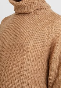 Sweter z golfem w jasnobrązowym kolorze, z teksturowanej dzianiny, o luźnym kroju i ściągaczu, który prezentuje ciepły i przytulny design.