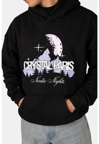 Černá mikina s kapucí zobrazuje grafiku hor a měsíce v purpurové a bílé barvě. Obsahuje text "CRYSTAL PARIS" a "Nordic Nights."