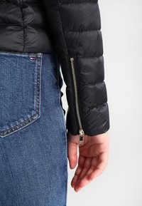 Ärmen på en svart vadderad jacka med quiltad struktur och en silverfärgad dragkedja som detalj. Matchas med blå jeans.