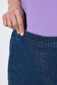 Personne portant un jean bleu à taille élastique et une chemise violette, tirant légèrement la ceinture du corps.