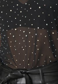 Svart mesh-topp med reflekterande rhinestones över materialet, med V-ringning och lagerdesign ovanför ett svart bälte.