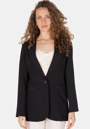 Donna con capelli ricci che indossa una giacca nera sopra una maglietta bianca con bordatura in pizzo, in piedi su uno sfondo chiaro e uniforme.