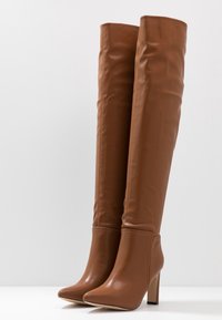 Botas sobre la rodilla de piel sintética marrón con punta cuadrada y tacón de aguja alto. Textura suave con un mínimo de detalles de costura.