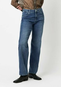 Blaue Jeans mit geradem Bein, fünf Taschen und Knopfverschluss. Getragen mit schwarzen Leder-Ankle-Boots.