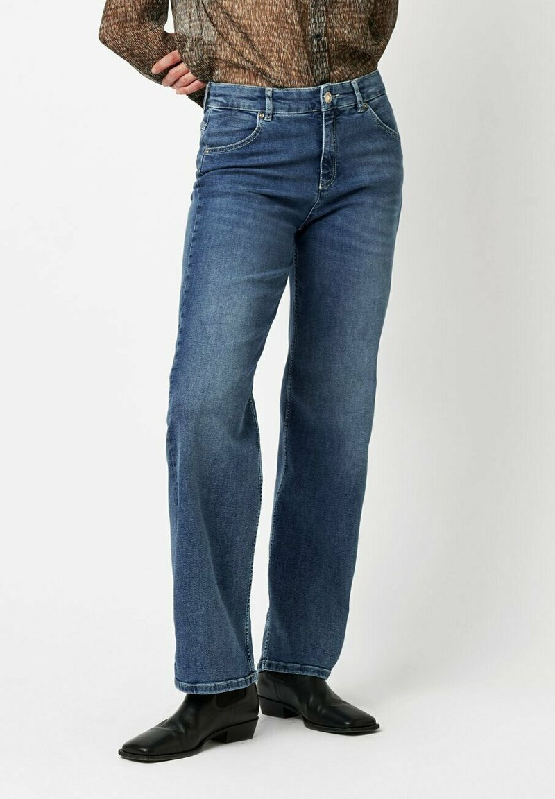 Blaue Jeans mit geradem Bein, fünf Taschen und Knopfverschluss. Getragen mit schwarzen Leder-Ankle-Boots.