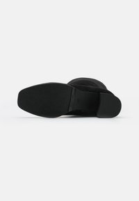 Bota de ante negra con suela plana y texturizada. El diseño presenta un empeine liso y un hardware mínimo, enfatizando una forma elegante y moderna.