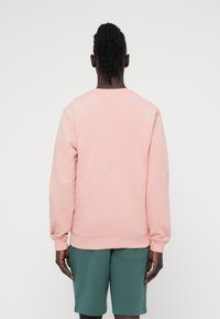 Ανοιχτό ροζ φούτερ με ριμπ μανσέτες και τελείωμα, φορεμένο πάνω από πράσινα σορτς. Διαθέτει απλό σχέδιο και ομαλή υφή υφάσματος.