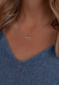 Sølv halskæde med "Aloha" tekst i kursiv skrifttype. Båret om halsen over en blød blå v-hals sweater med en pelsagtig tekstur.