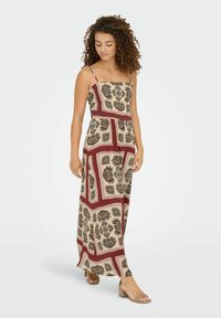 Femme aux cheveux bouclés portant une robe maxi à motifs floraux beige et bordeaux et des sandales à talons blocs beiges, se tenant contre un fond uni.